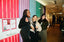 Three women smiling and posing together in a brightly lit indoor event space with colorful striped walls and a hallway in the background.