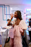 A woman in a white puff-sleeve top and a fluffy pink coat holds a white tote bag with the brand name OLFACTORY NYC while standing in a modern retail space.